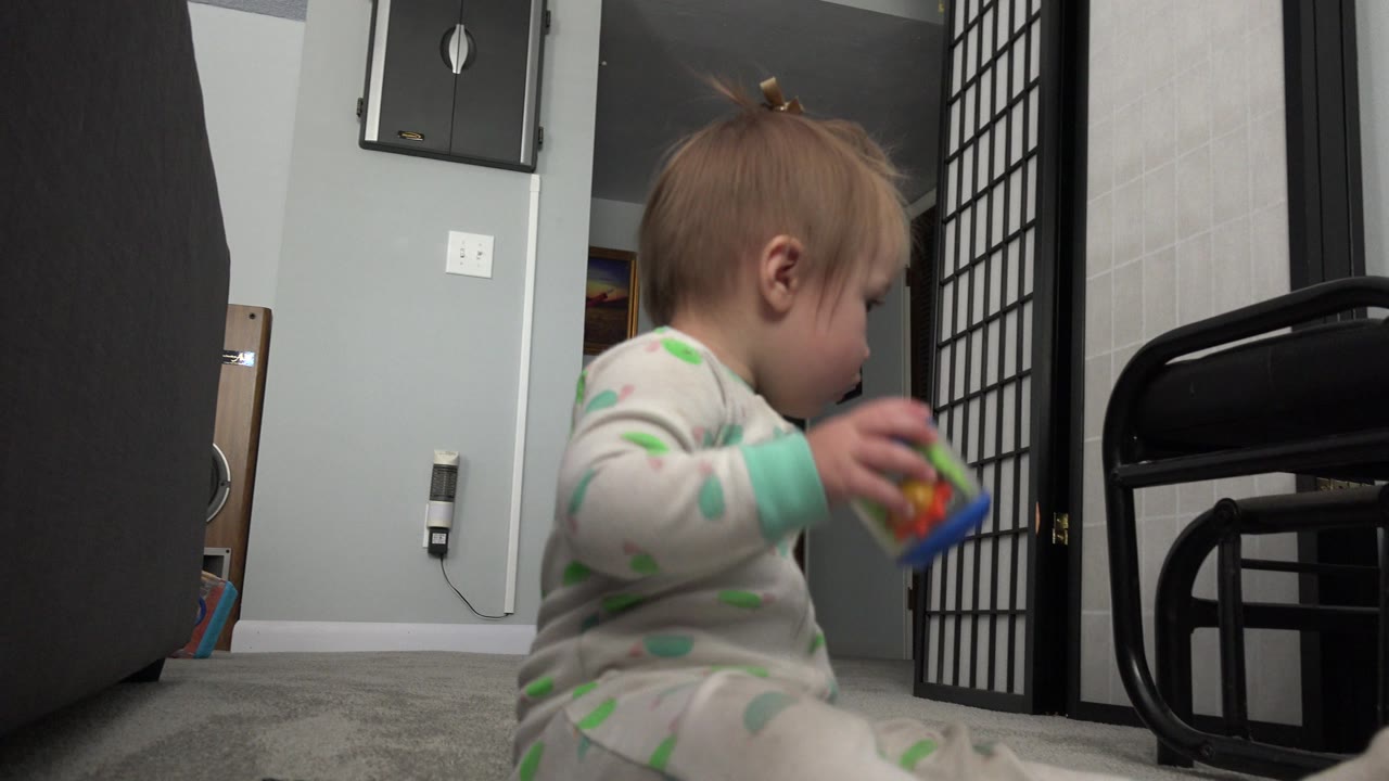 The toddler looks down at the toy while seated on the carpet, with the black divider and furniture framing the indoor home background.