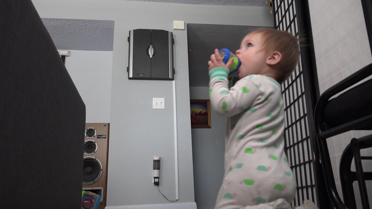 At 0:01 in Westminster, Colorado, the infant remains in profile with both hands around the small blue and green object, framed by dark foreground furniture and a black metal rail.