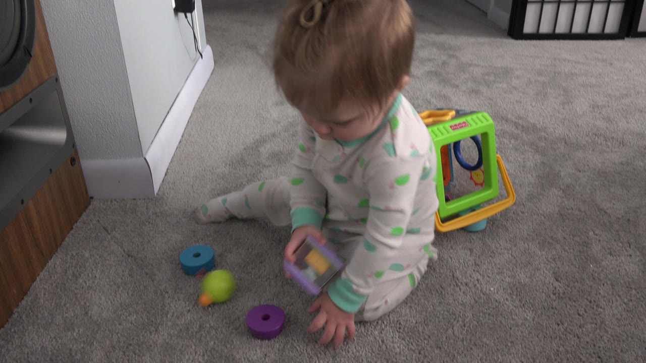 Blonde baby girl sitting on carpet with colorful toys nearby.
