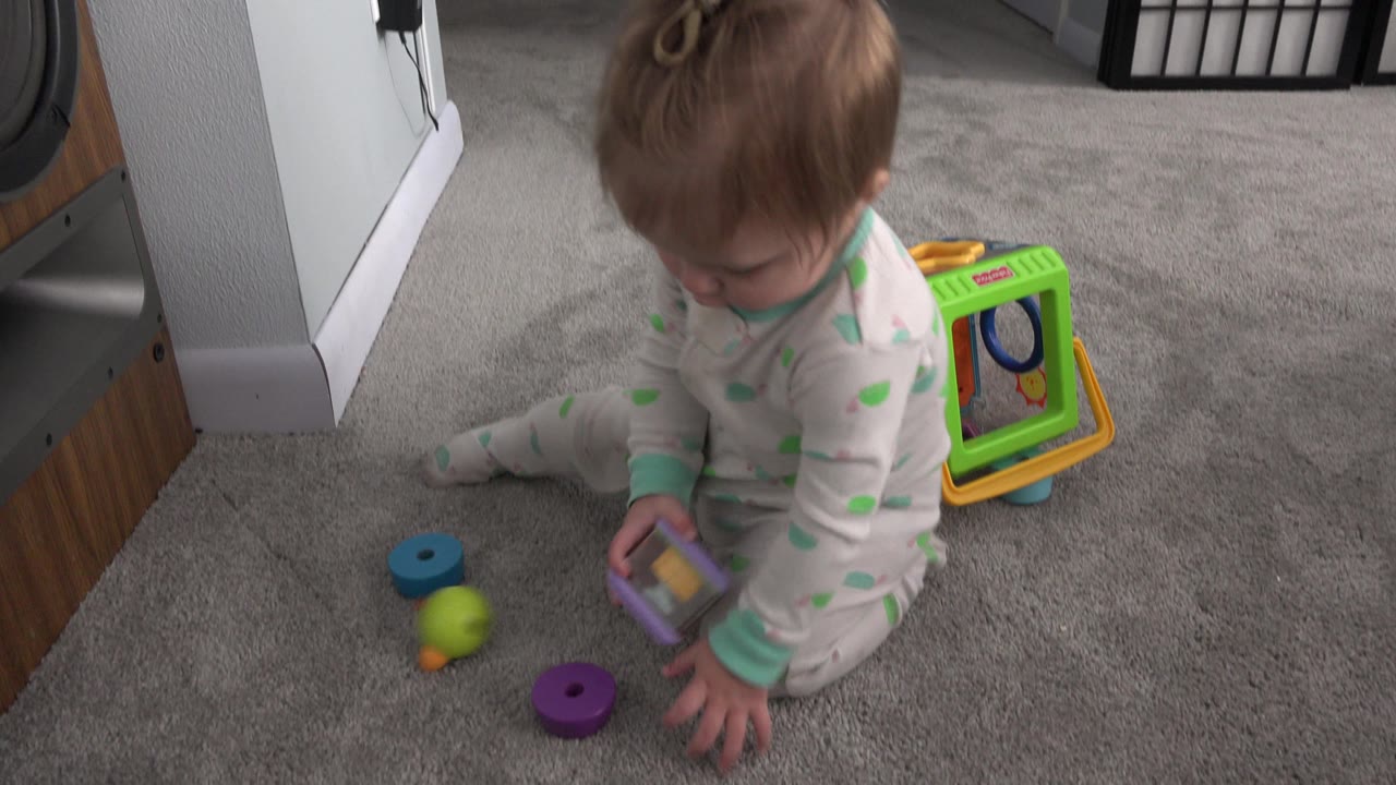 The baby girl remains seated on the carpet in the Westminster, Colorado interior, leaning toward a small purple and yellow toy while other bright pieces sit nearby.