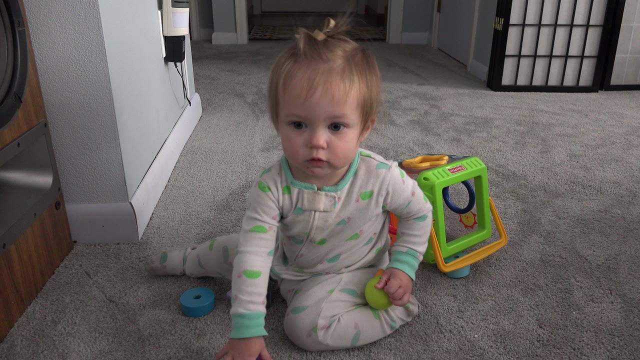 The child remains seated facing forward on the gray carpet, with a green toy cube behind one arm and a blue circular toy near the floor.
