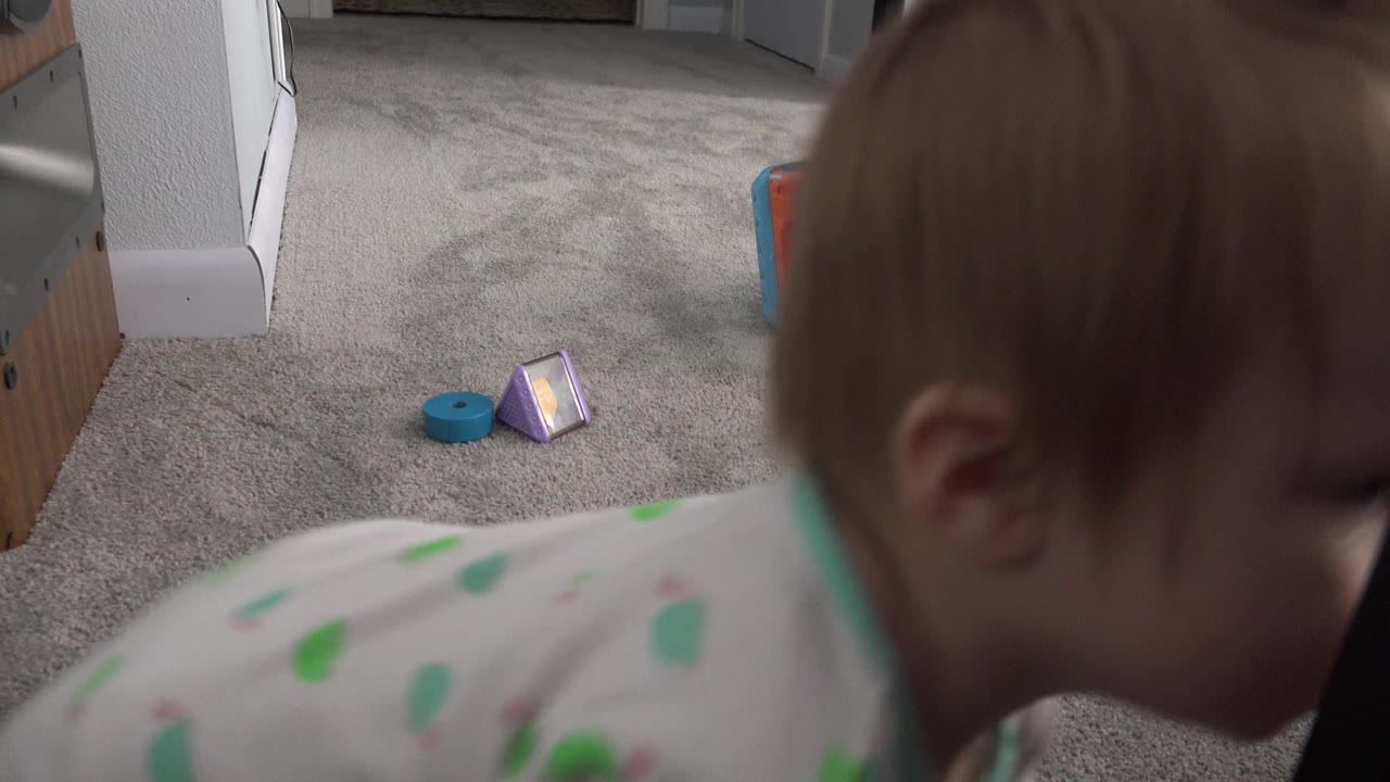 The toddler remains near the lens in the Westminster home interior, with his face mostly turned down and colorful toys still visible on the gray carpet in the background.