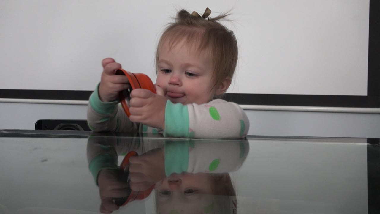 The baby keeps both hands on the black and orange object and looks downward, with the glossy tabletop reflecting the face and sleeves.
