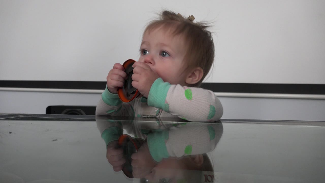 Baby holds an orange bell near their mouth behind a reflective tabletop.