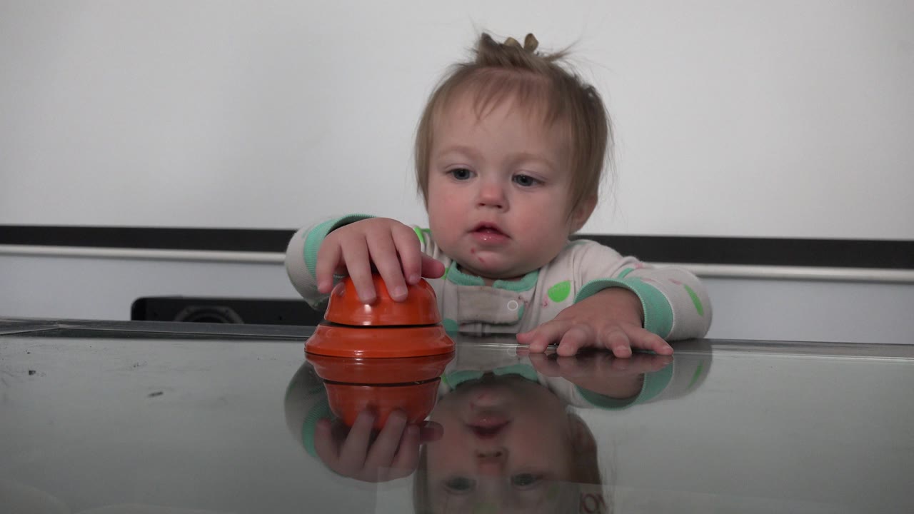 Toddler reaching toward a red hotel bell on a reflective tabletop.