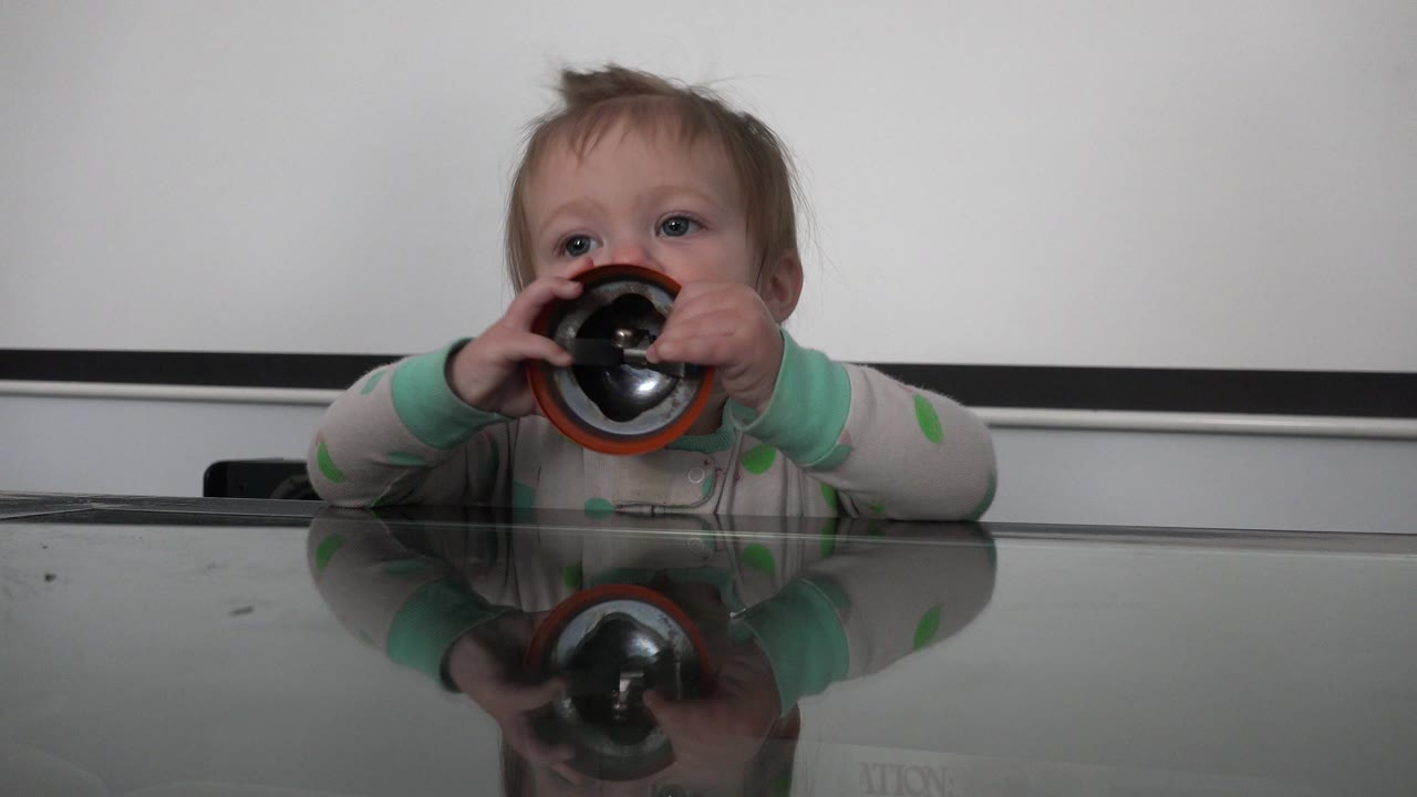 Blonde toddler holds a black and red object to their mouth at a glass table.