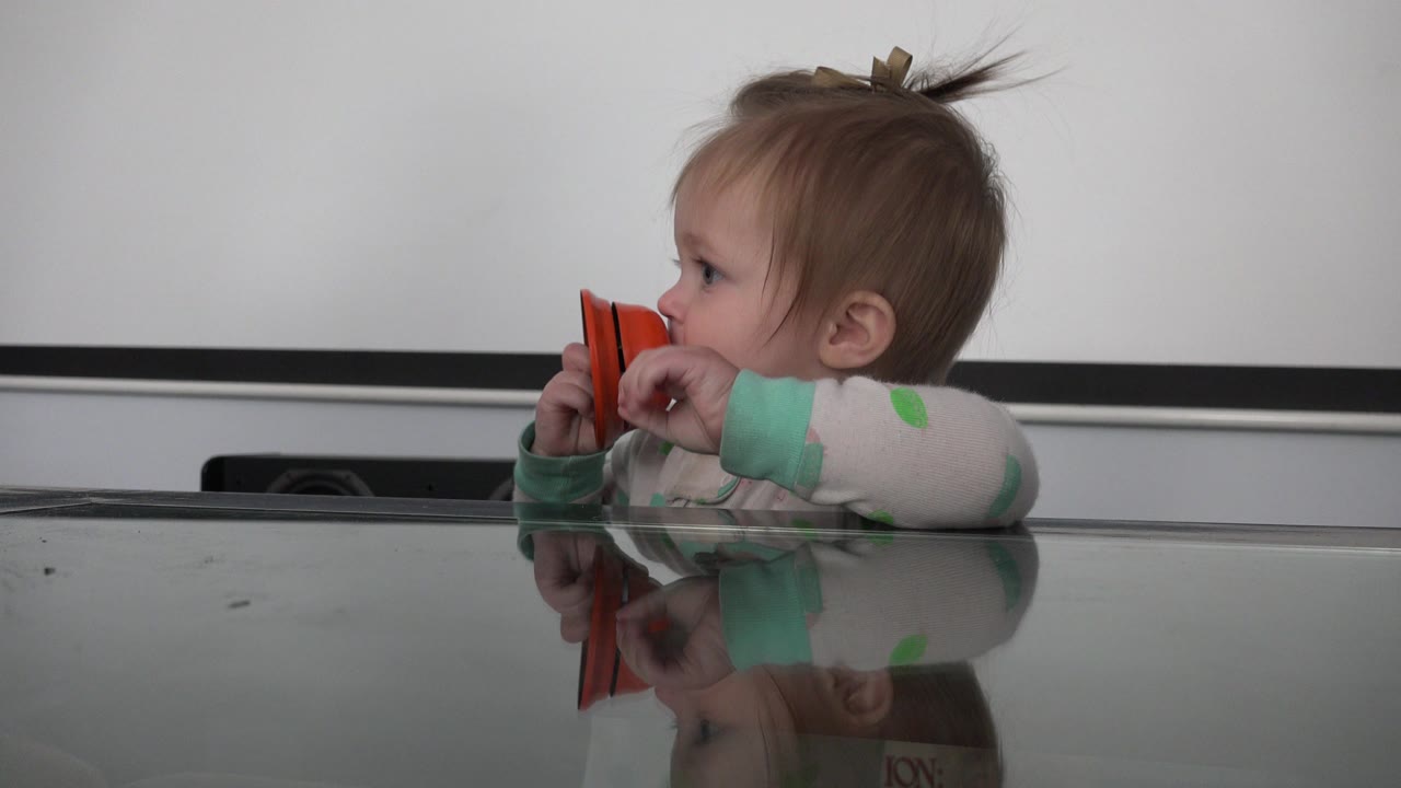 Baby holds an orange object near their mouth behind a glass table.