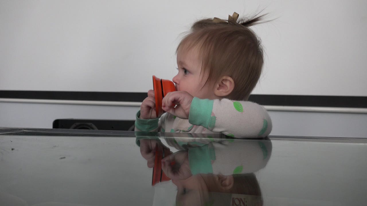 In Westminster, Colorado, the baby looks to the side while keeping the orange instrument near the mouth, with the glass tabletop reflecting the child and object.
