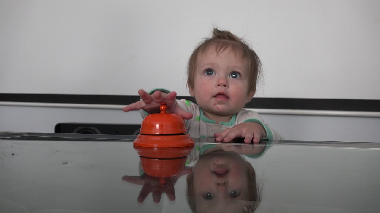 Toddler reaching toward an orange toy on a reflective table.