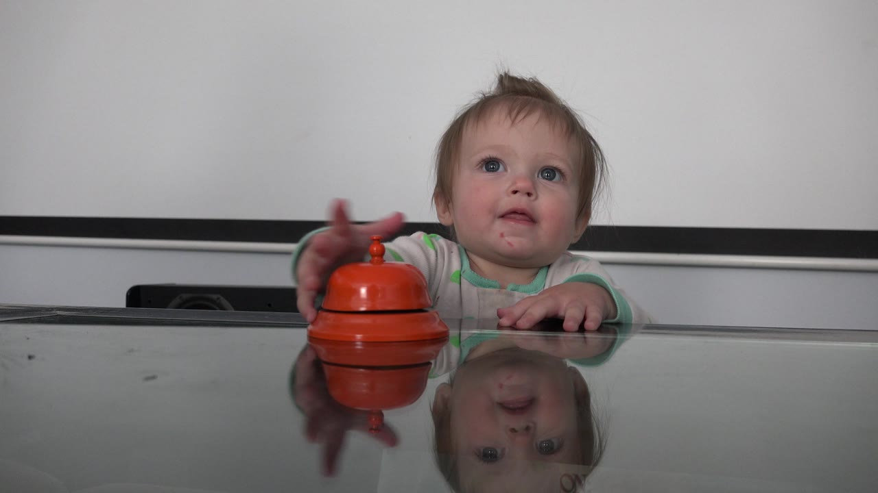 A toddler in Westminster, Colorado reaches over the glass tabletop and touches the top of the orange bell-like toy, with a faint smile and table reflection visible.