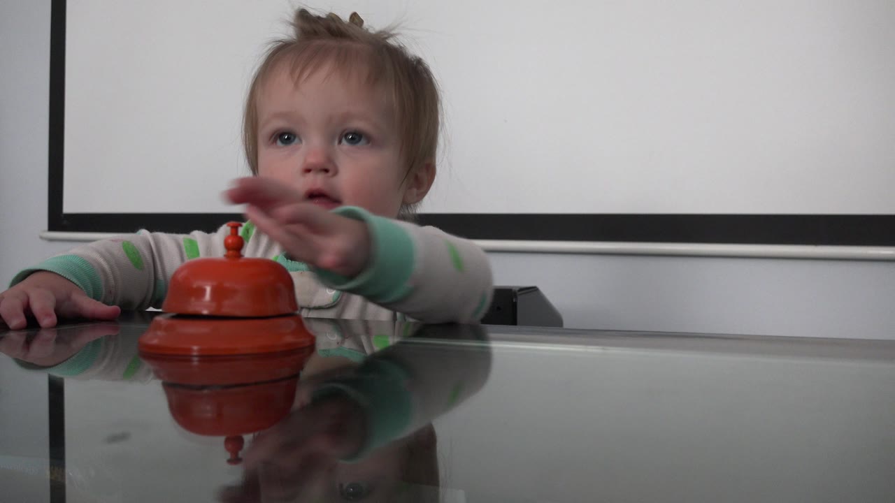 Toddler reaches toward an orange-red bell on a reflective table.