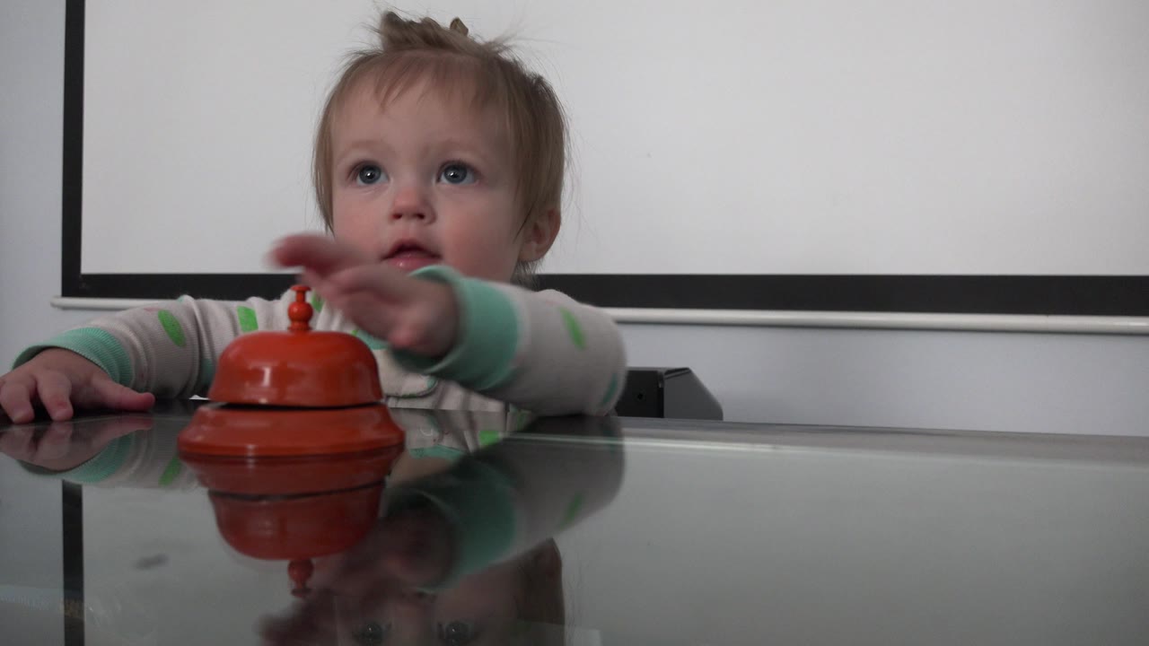 The child sits behind the reflective table in Westminster, Colorado, extending one hand toward the orange-red bell while looking slightly upward.