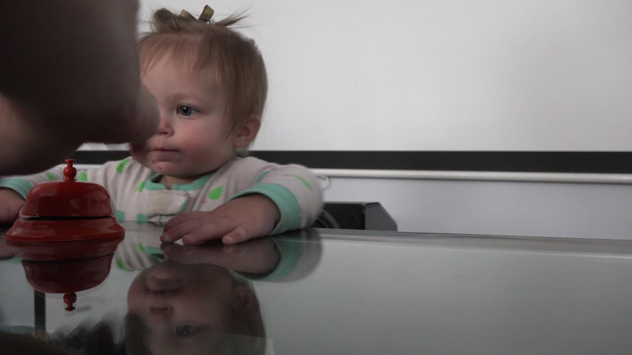 In Westminster, Colorado, an infant sits behind a reflective glass tabletop while a red bell-like object rests in front and an adult arm appears at the edge of the frame.
