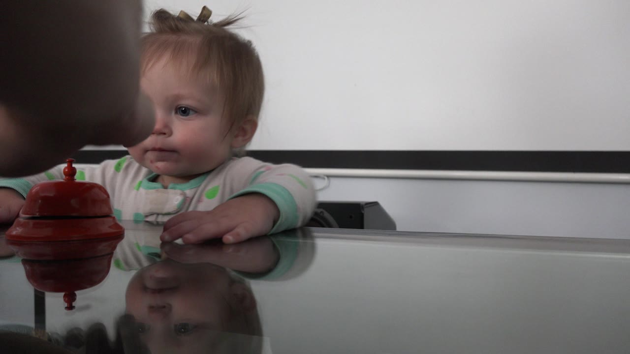In Westminster, Colorado, the infant looks toward an adult hand hovering near the small red bell-like object on a reflective glass table.