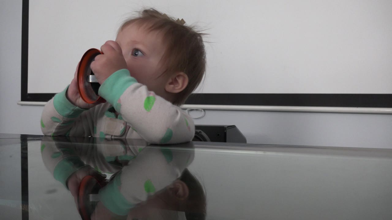 Baby holding an orange and black bell at a glass table.