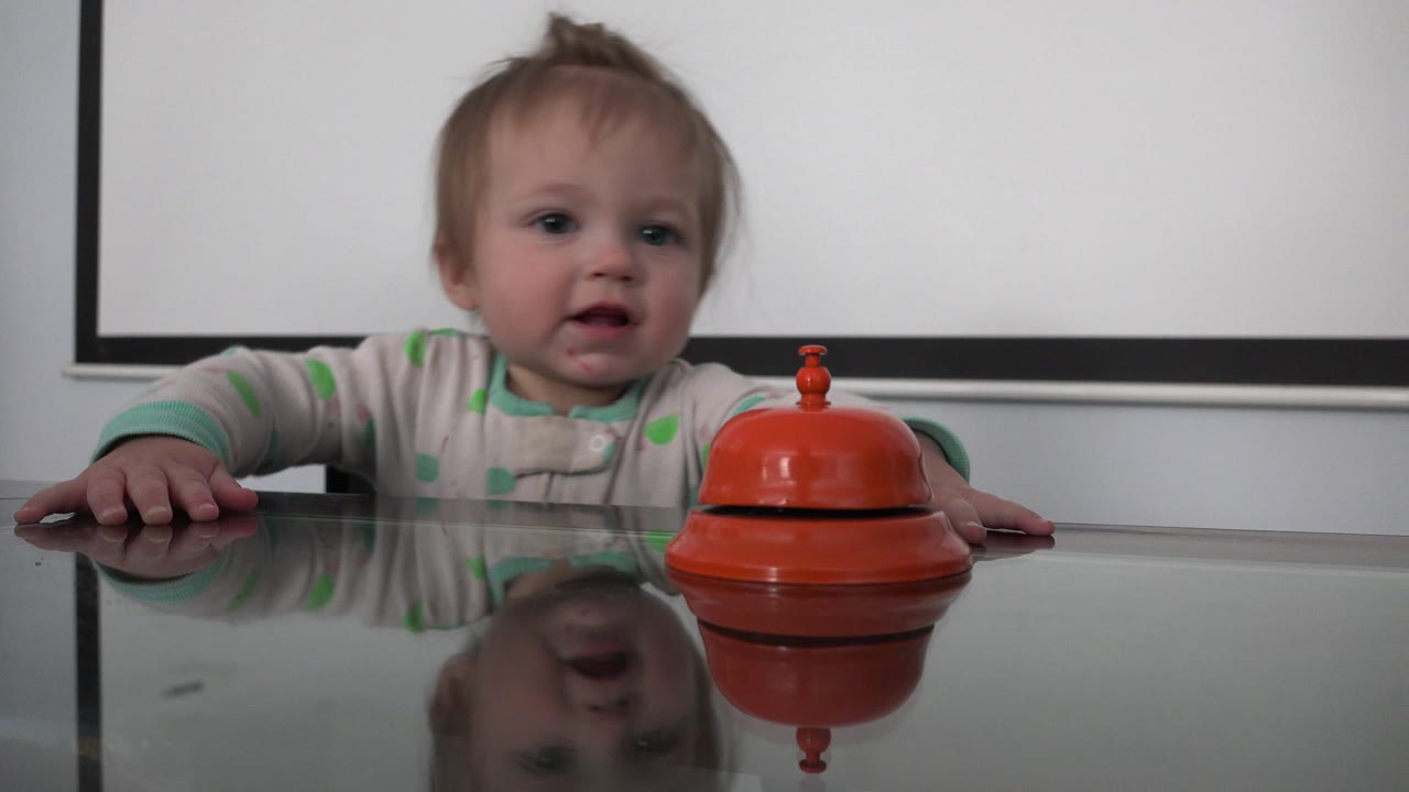 A toddler in Westminster, Colorado sits behind a reflective glass dining table with one hand on the surface and an orange toy centered nearby.