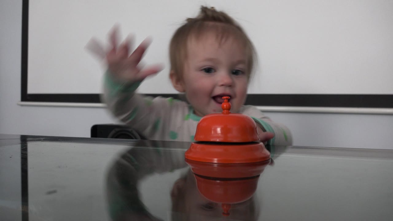 A toddler in Westminster, Colorado lifts one hand in motion beside the orange toy while seated at the reflective glass dining table.