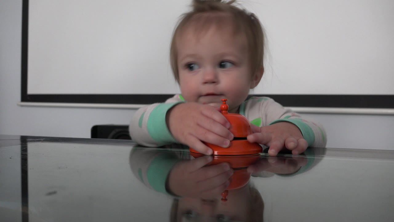 At 0:01 in Westminster, Colorado, the child keeps one hand on the orange object and glances sideways above the reflective glass table.