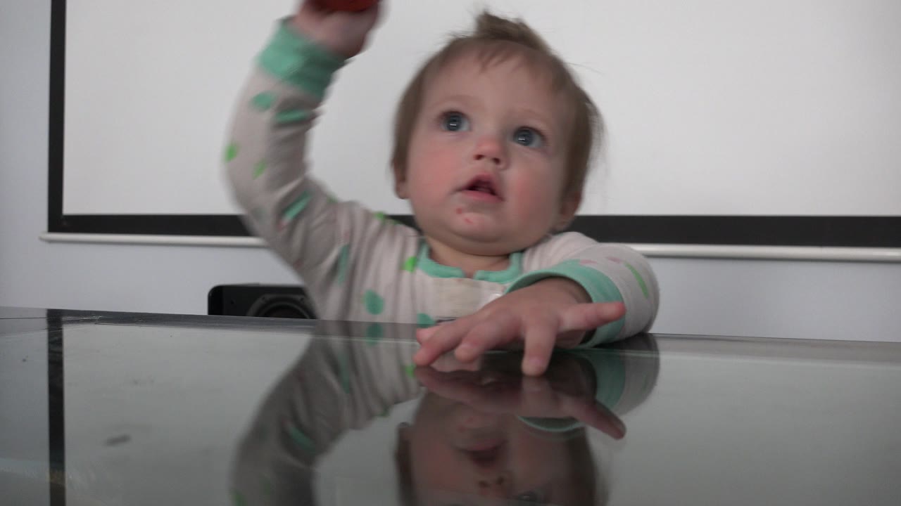 A toddler in light pajamas sits at a reflective glass dining table in a Westminster, Colorado home interior, looking up while one arm reaches above the frame.