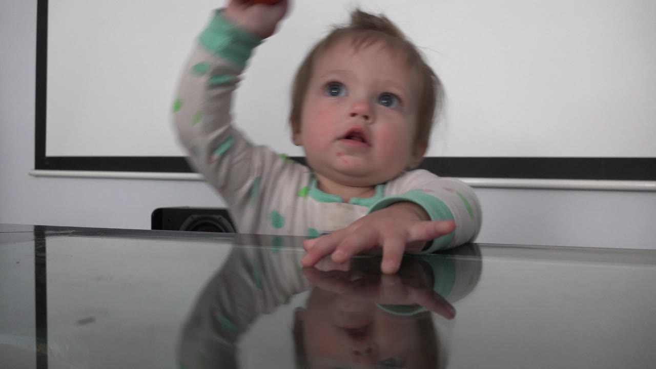 The toddler remains centered behind the glass tabletop in the Westminster home, eyes raised and one hand lifted above the visible frame.