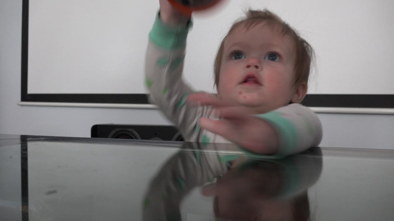 A toddler at the reflective dining table reaches with both hands as an orange and black round object enters near the top of the frame in the Westminster interior.