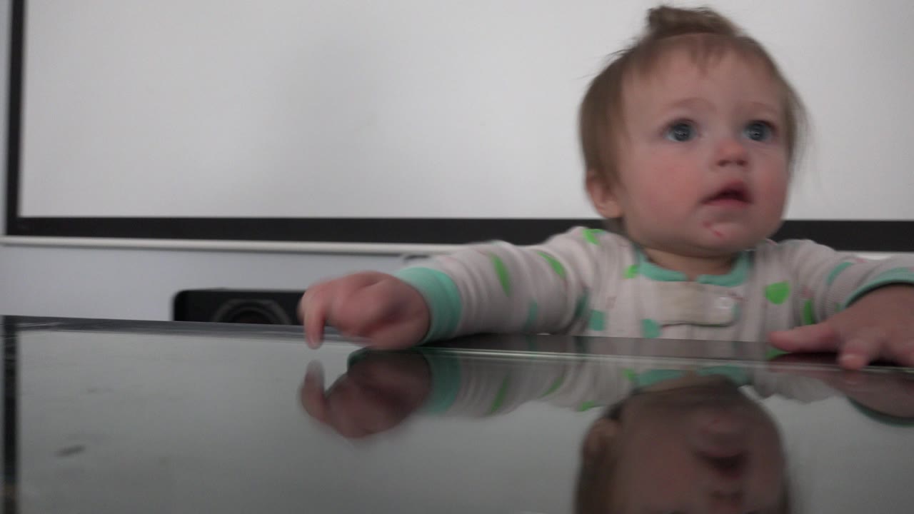 A baby in striped pajamas stands behind a reflective glass table in Westminster, Colorado, looking upward with both hands near the tabletop.
