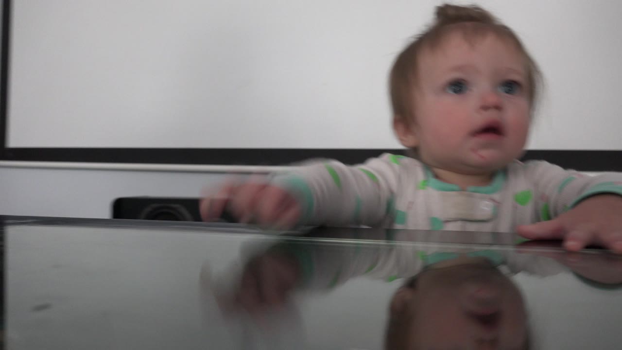 The baby remains centered behind the glass table in Westminster, Colorado, with one hand moving forward and a soft reflection visible on the tabletop.