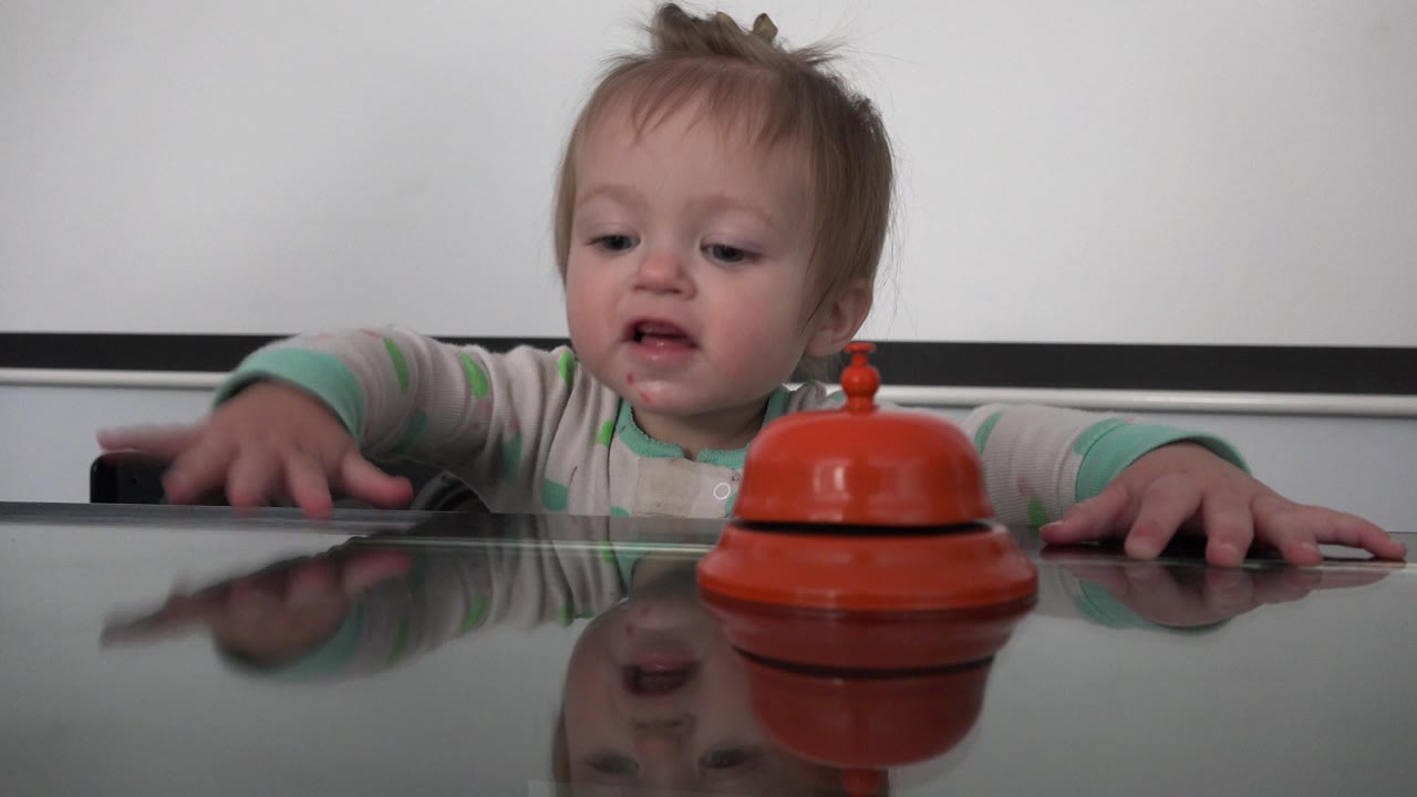 Blond child reaching near an orange bell on a glass table.