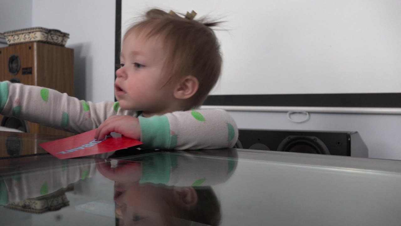 The toddler looks to the left with an arm extended over the glass table, while the red paper item rests near their other hand in the Westminster interior.