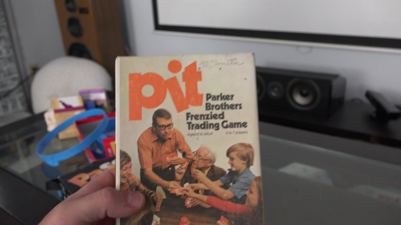 A hand holds a Pit Parker Brothers Frenzied Trading Game box in a Westminster, Colorado home interior, with toys, speakers, and a lamp behind it.