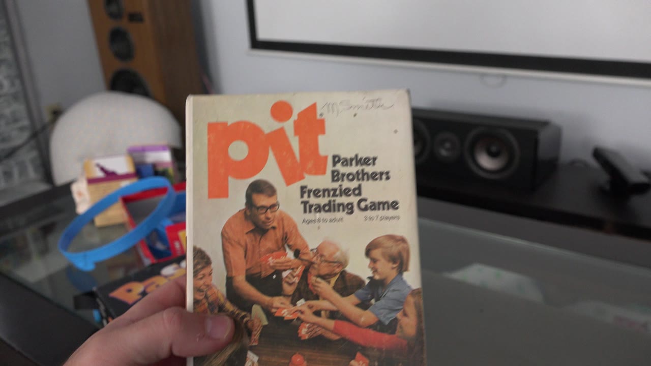 The Pit game box remains held close to camera in the Westminster, Colorado home, with the glass tabletop and dark speakers still visible in the background.