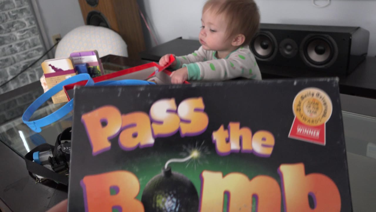 A baby girl in a Westminster, Colorado home sits behind a large Pass the Bomb board game box on a glass table, with speakers and a brick-pattern wall in the background.