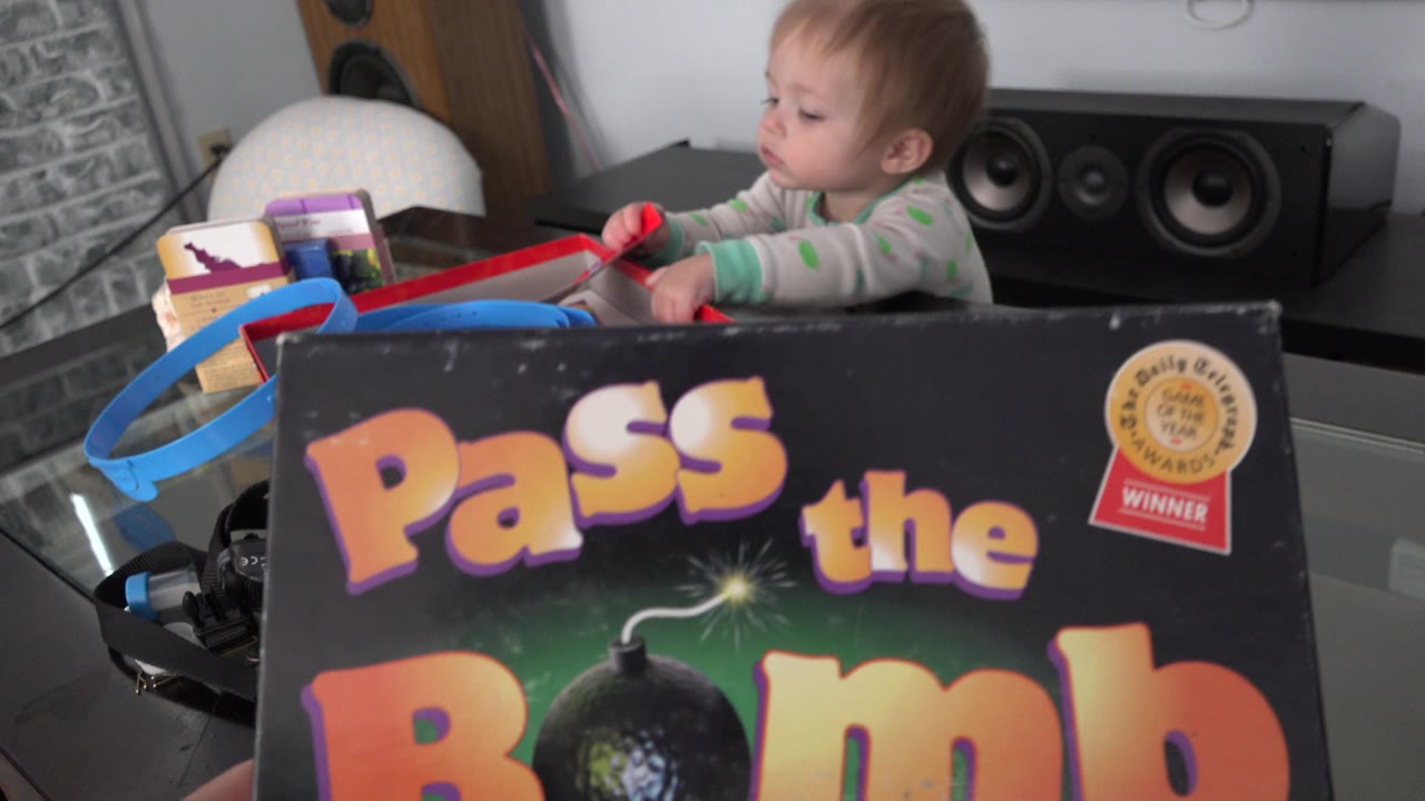 A baby girl in a Westminster, Colorado interior reaches near a red game piece while the Pass the Bomb box fills the lower foreground and audio speakers sit behind her.