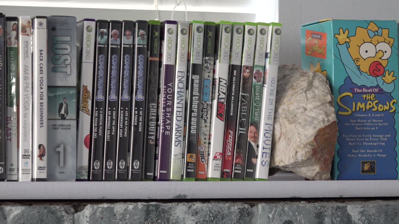 A shelf in Westminster, Colorado shows rows of Blu-ray and DVD cases, a large rock, bright window blinds, and a Simpsons-branded box, with no visible people.