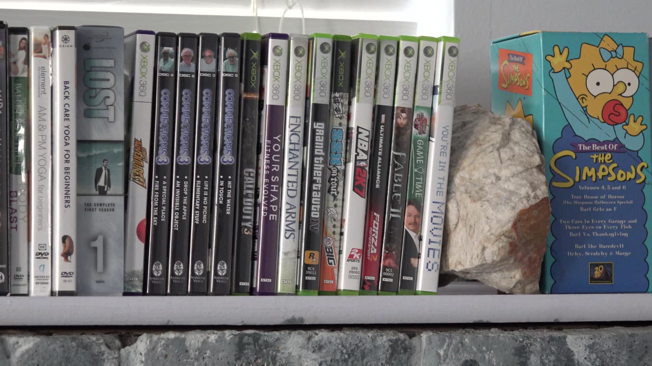 The Westminster interior shelf remains centered on movie cases, a pale rock, and the Simpsons-branded box against backlit blinds, with no visible people.