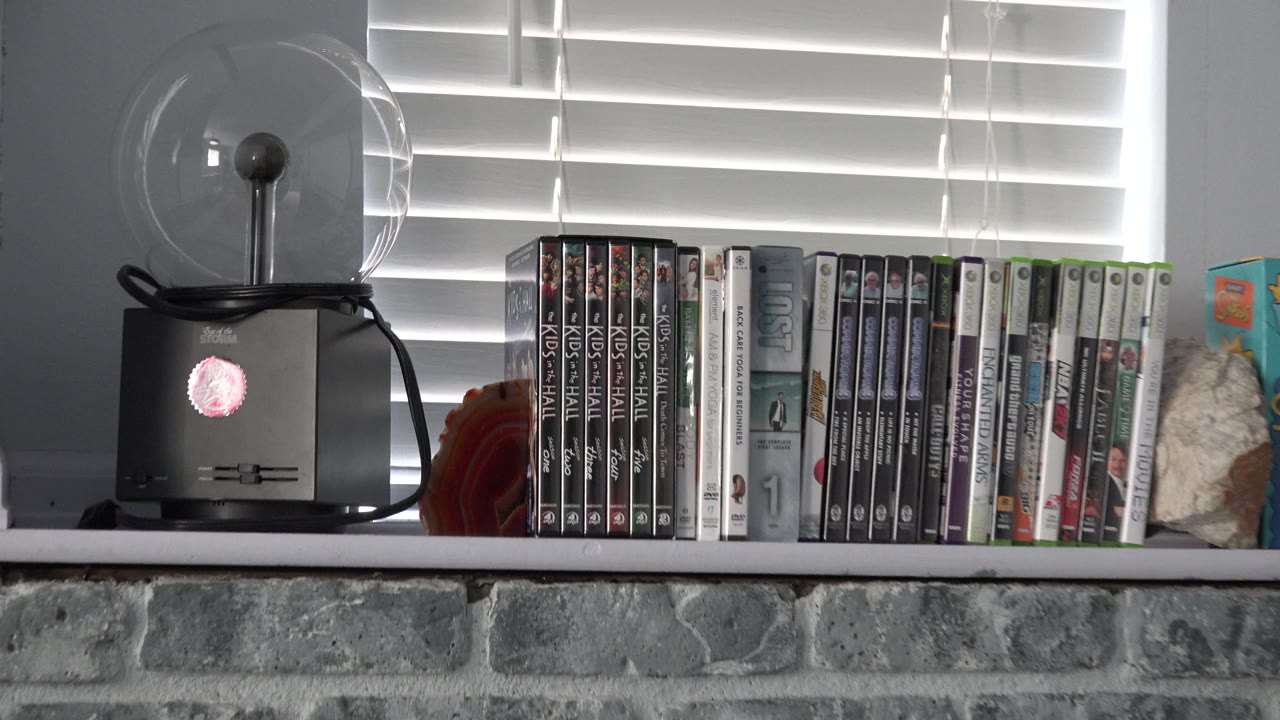 Westminster, Colorado home interior showing a shelf of video game cases beside a small fan and white window blinds above a gray stone fireplace, with no visible people.