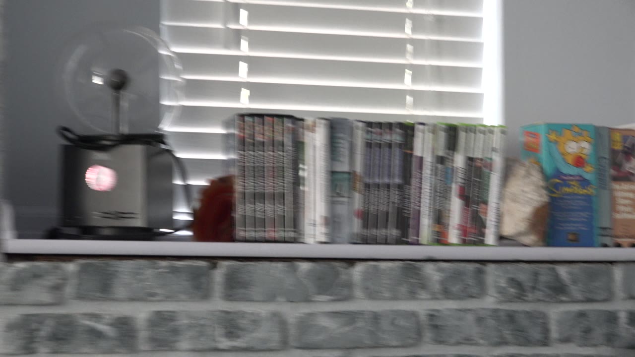 Blurred shelf of game cases beside blinds and a small fan.