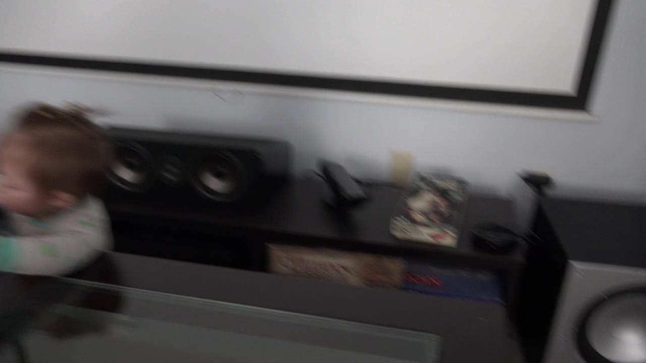 A child is partly visible in motion blur near the left side of a black entertainment center with stereo speakers and boxed games in a Westminster, Colorado home.