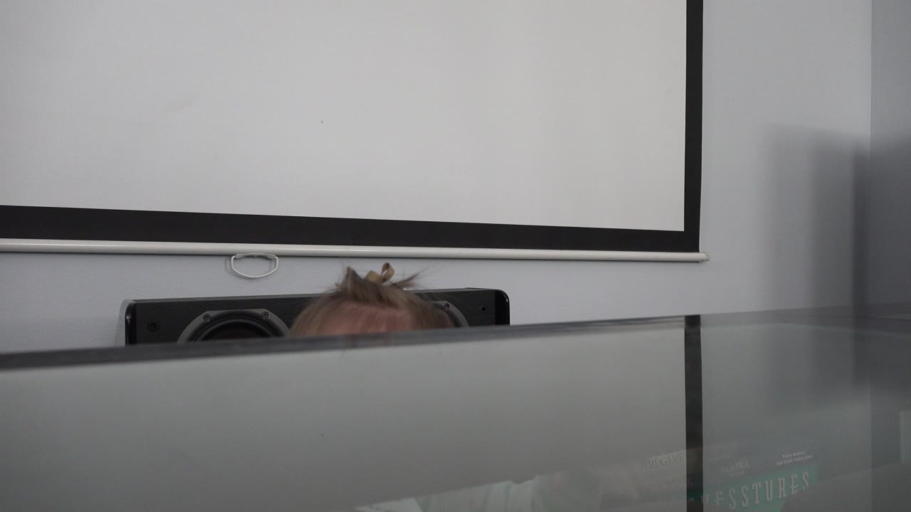 At 0:00, the top of a baby girl's head just appears behind a reflective glass table in a Westminster, Colorado home interior, with a white pull-down screen on the wall behind her.