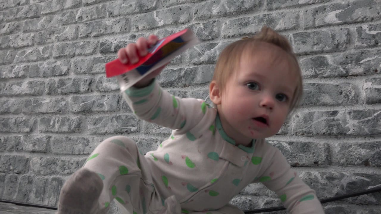Baby on the floor holding a red object near a gray brick-pattern wall.