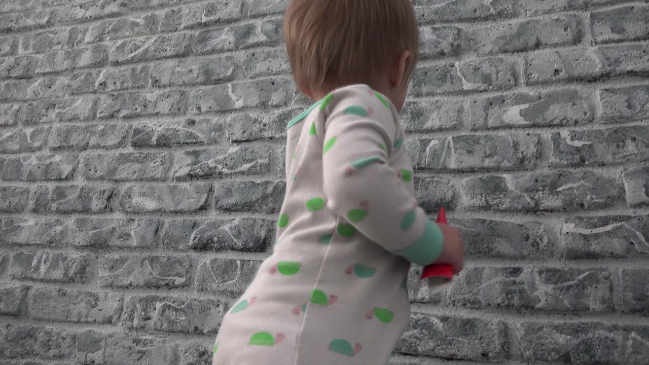 At 0:01 in Westminster, Colorado, the toddler remains near the grey brick wall, turned slightly to the side with the red object partly visible near the right hand.