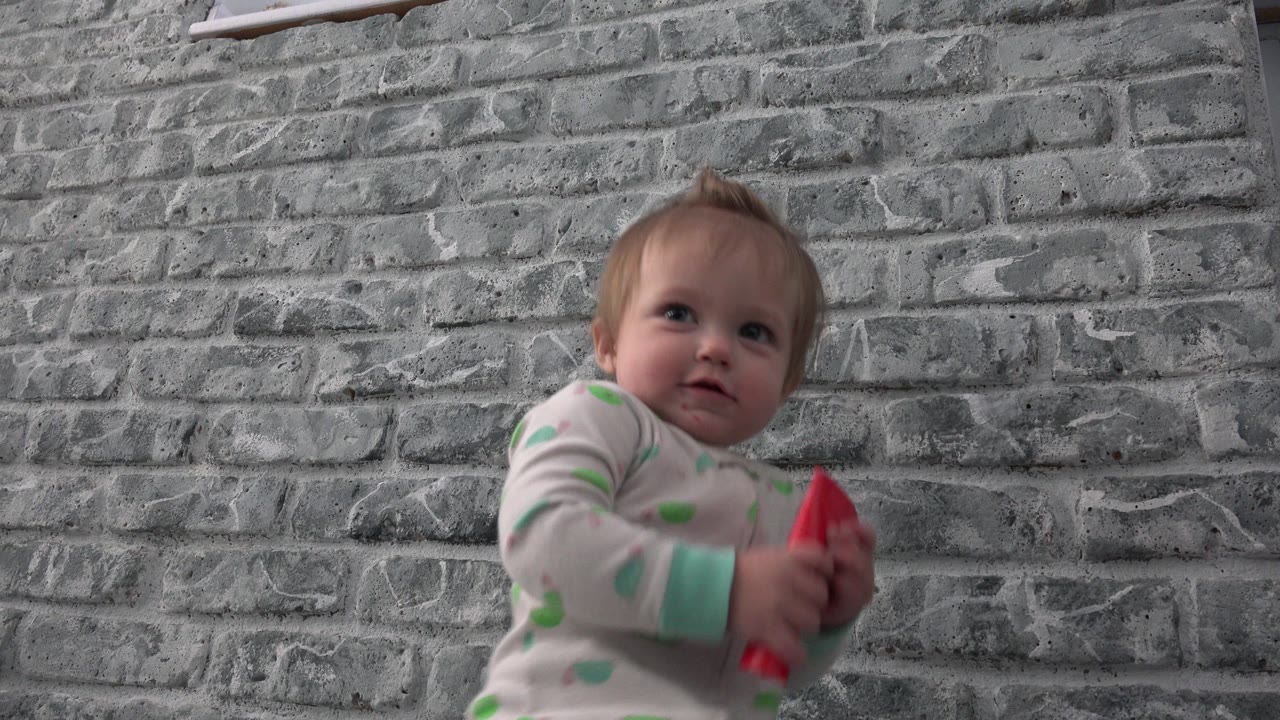 Baby in pajamas holding a red card in front of a gray brick wall.