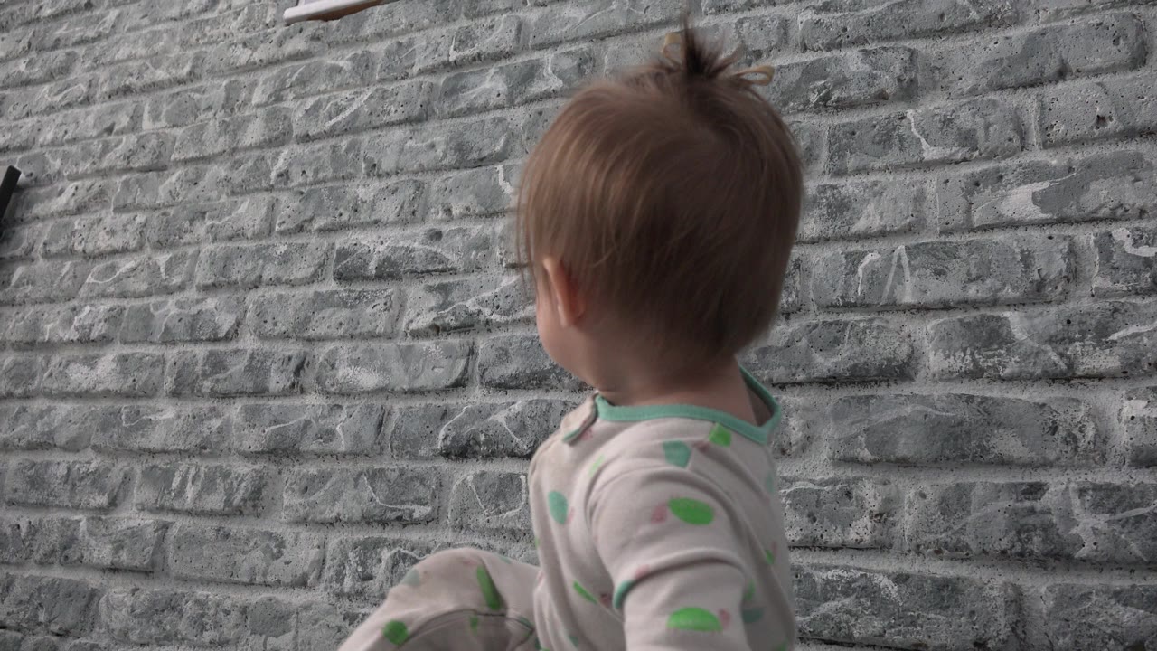 Young child seated beside a gray brick wall, turned away from camera.