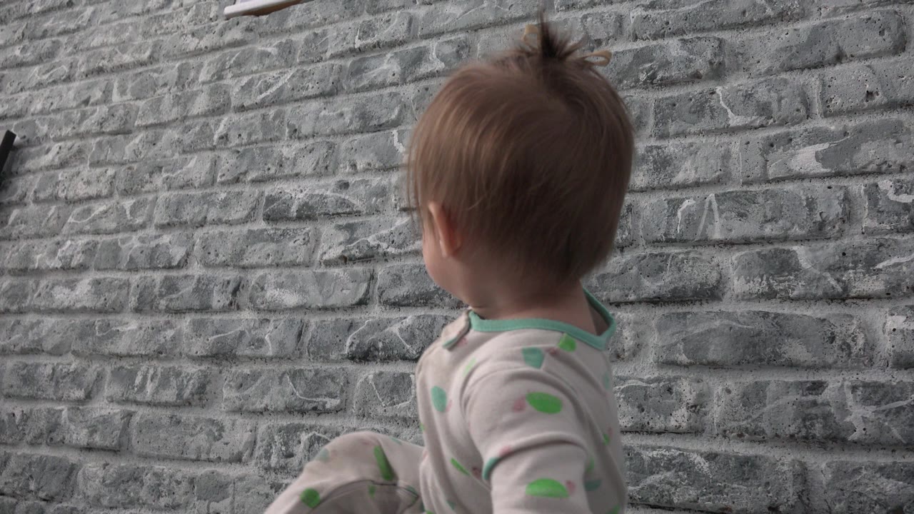 At 0:01, the child remains seated by the gray brick wall in Westminster, Colorado, with a slight turn of the head showing more of the side profile.