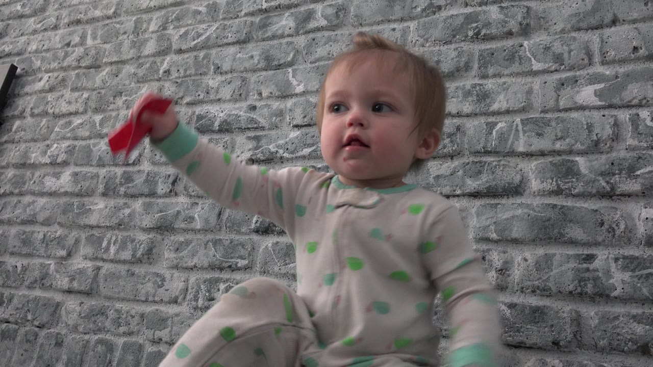 Toddler in a patterned sleeper raises a red object indoors.