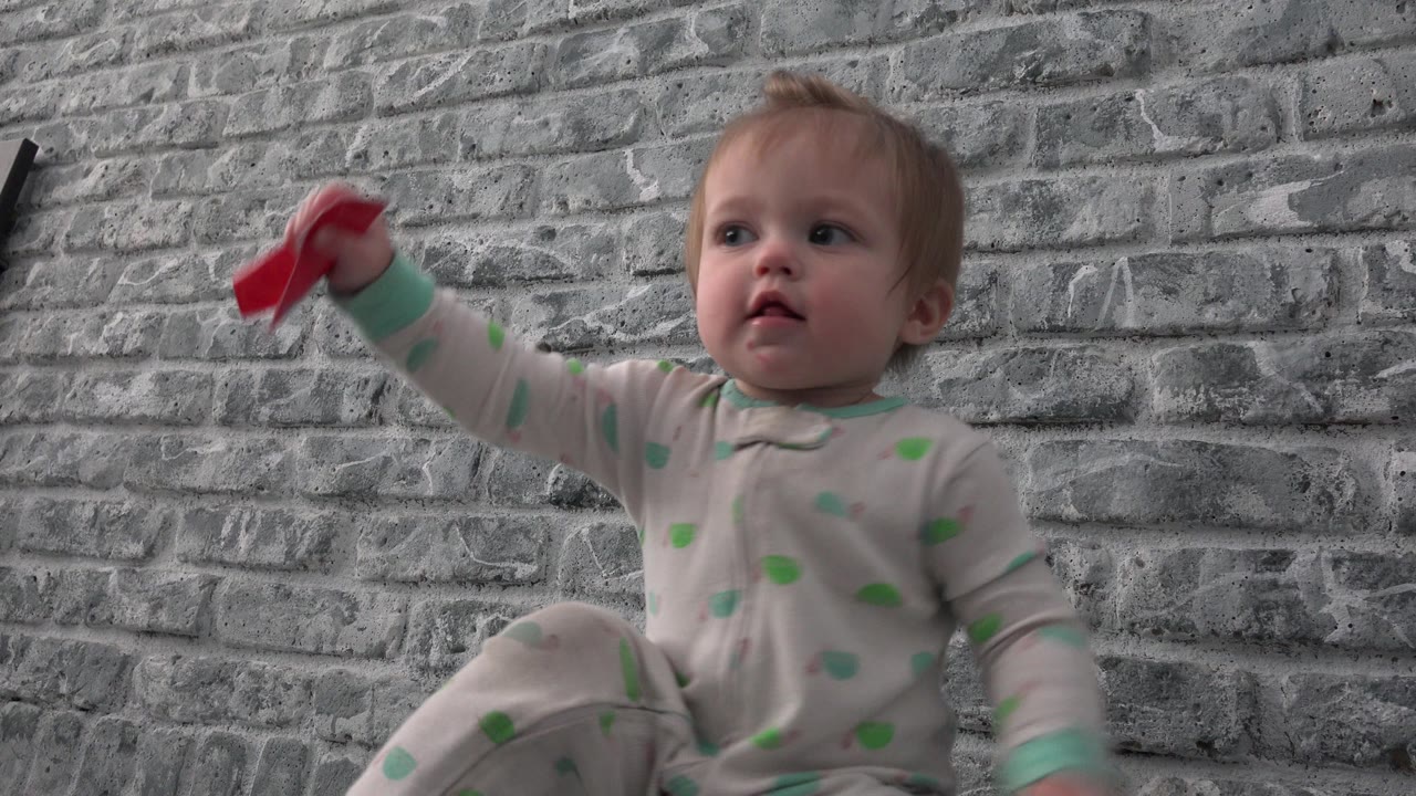 The toddler remains seated indoors in Westminster, Colorado, holding the red object outward while looking slightly off camera.