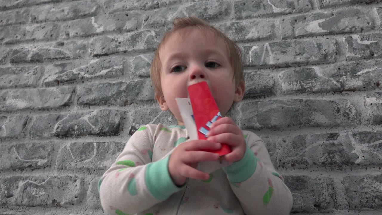 At 0:00 in Westminster, Colorado, a baby in pale pajamas sits low against a gray brick-pattern wall while holding a red-and-white paper toy near the mouth.
