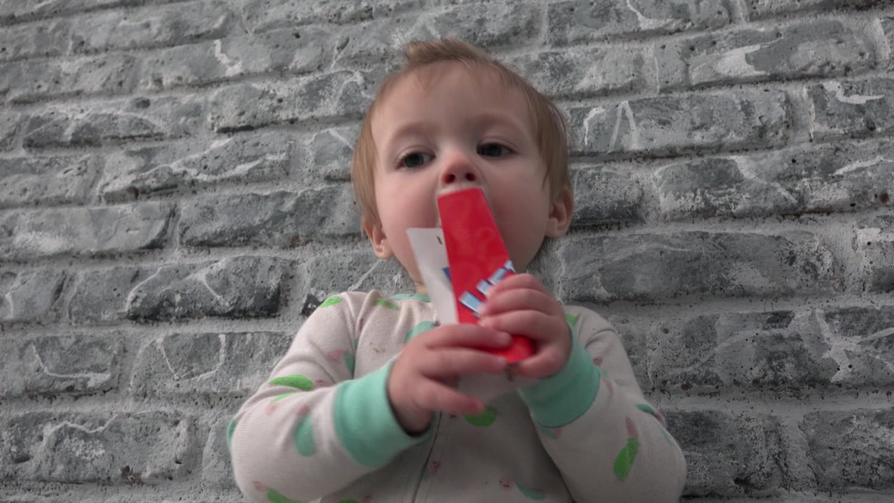 At 0:01 in Westminster, Colorado, the baby remains centered and seated, looking toward the camera while pressing the red toy to the mouth.