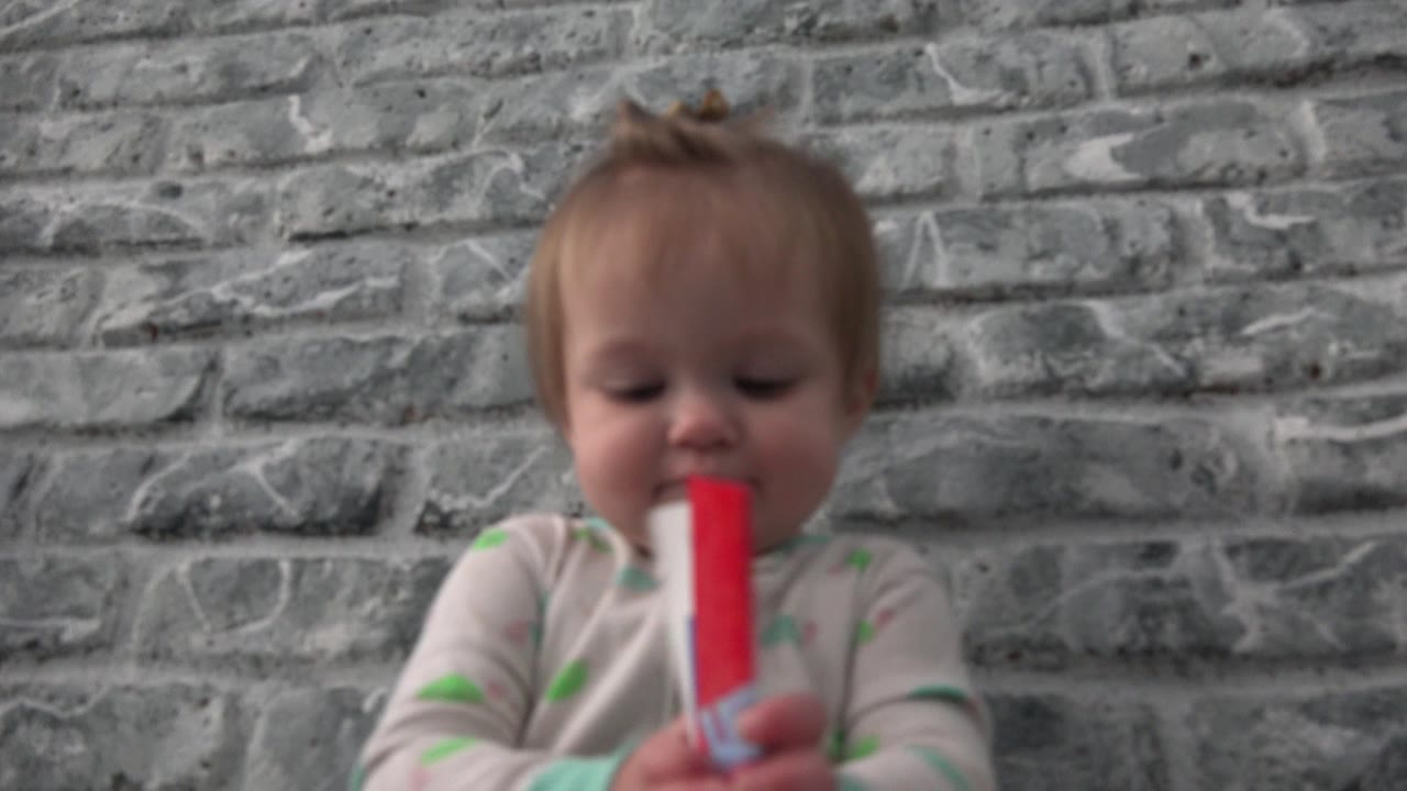 At 0:00, a baby in light pajamas holds a small red paper close to their mouth in Westminster, Colorado, with a gray brick-pattern wall filling the background.
