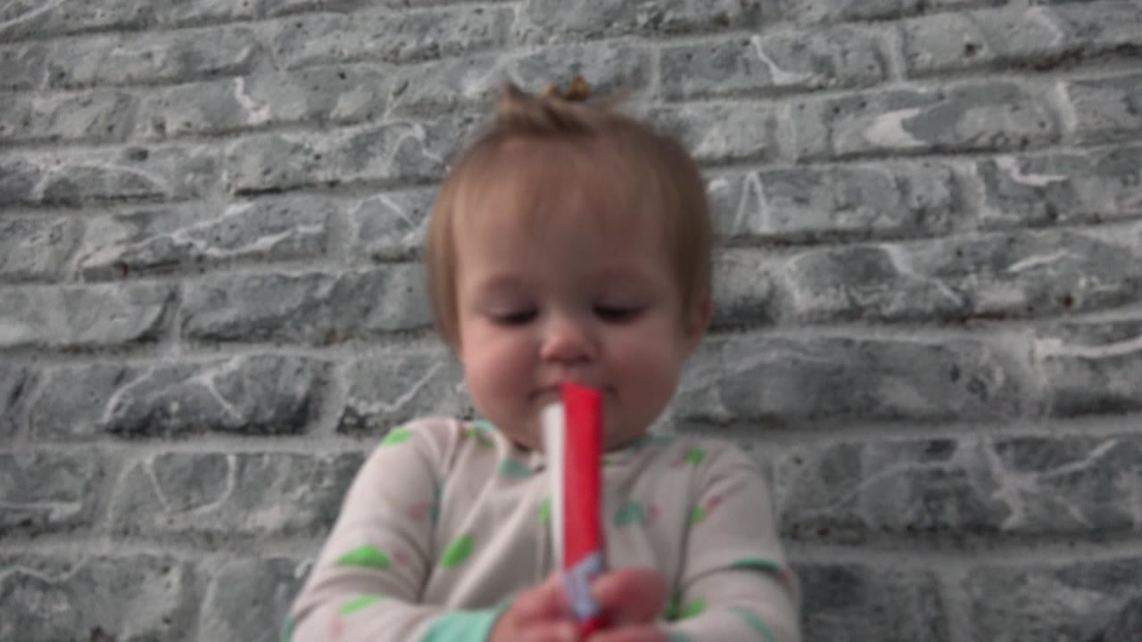 At 0:01, the baby keeps the red paper upright in both hands and looks down toward it, with the textured gray wall behind them in Westminster, Colorado.